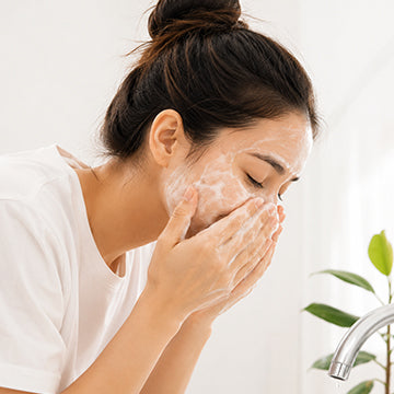 A woman with her hair in a bun washes her face with foaming cleanser at a sink, wearing a white t-shirt. A small green plant and a chrome faucet are visible in the bright background.